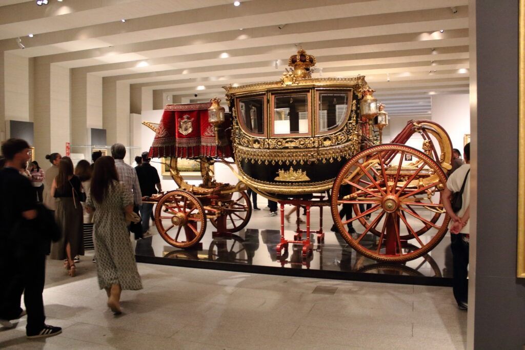 Un gran carruaje de gala ricamente decorado en negro y oro, con grandes ruedas rojas, expuesto en una sala de museo moderna de techos acanalados y luminosos.
