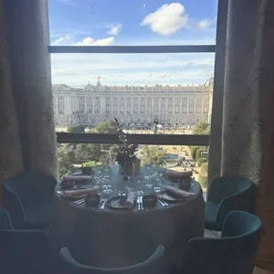 Mesa redonda elegante reservada para cenar junto a un ventanal alto con vistas panorámicas al Palacio Real de Madrid en un día soleado.