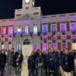 Groupe de personnes avec des badges posant de nuit devant la Casa de Correos à la Puerta del Sol, illuminée de lumières colorées.