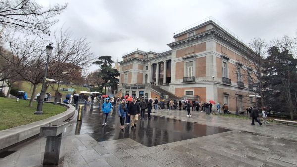 Acceso principal al Museo del Prado con visitantes haciendo cola en un día lluvioso en Madrid.