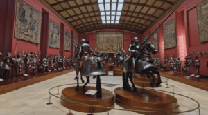 Vista de la sala principal de una armería real con caballeros en armadura montados a caballo sobre pedestales, rodeados de tapices históricos.