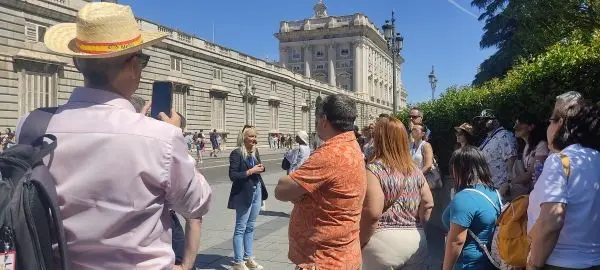 Un grupo de turistas escucha a una guía frente a la fachada lateral del Palacio Real de Madrid en un día soleado.